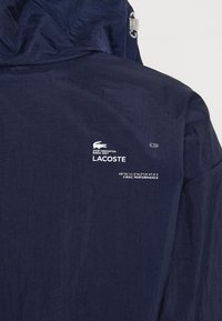 Námořnická modrá lehká bunda s texturovaným povrchem. Obsahuje kapuci, boční detaily a branding Lacoste s výkonnostními souřadnicemi.