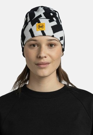 Black and white patterned knitted beanie with a yellow Buff logo patch. Fitted shape, made of soft fabric, worn by a model in a black sweater.