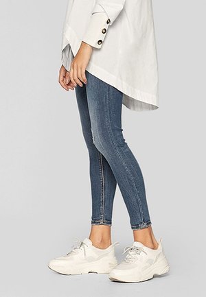 Person trägt blaue enge Jeans, weiße klobige Sneakers und ein weißes langärmeliges Hemd mit geknöpften Manschetten, steht seitlich.