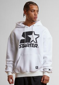 Hvid hoodie med et stort sort "STARTER" logo og stjerne grafik. Indeholder en frontlomme, snoretræk i huen og ribbet manchetter og kant.