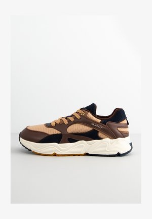 Light brown athletic sneaker with a mesh upper, dark blue accents, beige sole, and branded side detail. Rounded shape with textured panels.