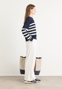 Sweater listrado em azul-marinho e branco, calças largas brancas e sapatilhas pretas. Bolsa ampla com detalhes em bege, azul-marinho e castanho.