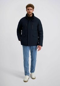 Mann in einer marineblauen Jacke mit Reißverschluss und Klapptaschen, hellblauen Jeans und weißen Sneakers, der vor einem schlichten hellen Hintergrund steht.