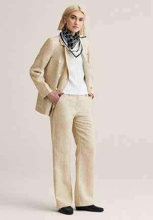 Kvinde iført beige jakkesæt med hvid striktop, sort mønstret halstørklæde og sorte loafers, stående mod en ensfarvet baggrund.