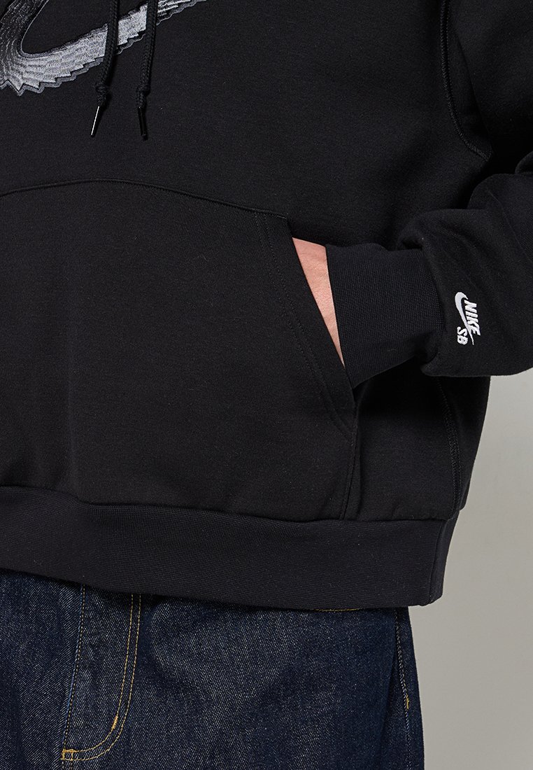 Hoodie noir avec un grand logo Nike gris. Il est doté d'une poche avant et de poignets côtelés. Porté avec un jean en denim foncé.