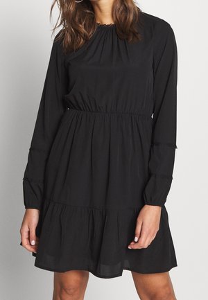 Mujer con vestido negro de manga larga con cintura fruncida y falda de capas, de pie frente a un fondo liso y claro.