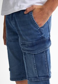 Džínové cargo šortky v tmavě modré barvě s několika kapsami, volným střihem a viditelnými švy. Látka vykazuje jemnou texturu.