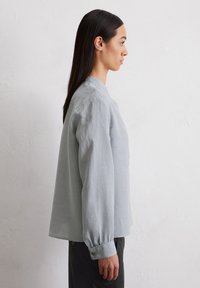 Chemise légère gris clair à manches longues avec une coupe décontractée, un col rond et des poignets boutonnés, fabriquée dans un tissu doux et texturé.