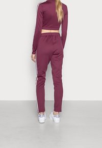 Completo sportivo color marrone scuro con top a maniche lunghe cropped e pantaloni affusolati, caratterizzato da strisce laterali bianche e una vestibilità rilassata, abbinato a sneakers bianche.