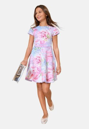 Nuori tyttö kävelee pastellinvärisessä kukkamekossa, pitää kädessään metallista laukkuetta, käyttää ballerinakenkiä ja rannekorua, hymyilee kevyesti sivulle.