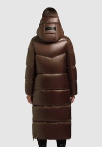 Hosszú barna puffertakaró, párnázott kialakítással, nagyméretű kapucnival és logóval a hátsó részén. Simagla, fényes anyag, függőleges és vízszintes varrásokkal.