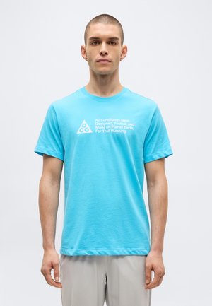 Homme portant un t-shirt turquoise avec un logo blanc ACG et un texte à propos de la course en sentier, debout devant un fond blanc uni.
