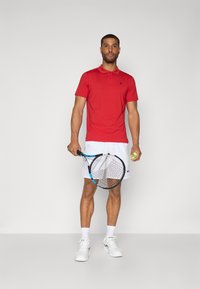 Polo rouge, shorts blancs, tenant une raquette de tennis et une balle. La chemise est à col, et les shorts arborent un logo. Des baskets blanches sont portées.