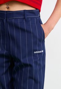Jūras zilas svītrainas bikses ar pielāgotu piegriezumu, ar baltām šuvēm un adidas logotipu kreisajā pusē blakus kabatai.