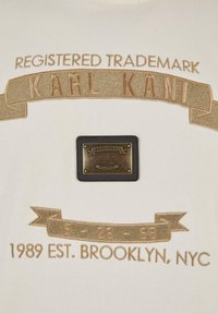 Krém színű textil arany hímzéssel, amelyen a "KARL KANI" felirat és olyan részletek találhatók, mint egy téglalap alakú bronz folt és egy banner dizájn.