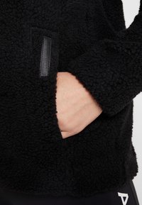 Main insérée dans la poche latérale d'une veste polaire noire texturée avec une étiquette verticale « Didriksons ».