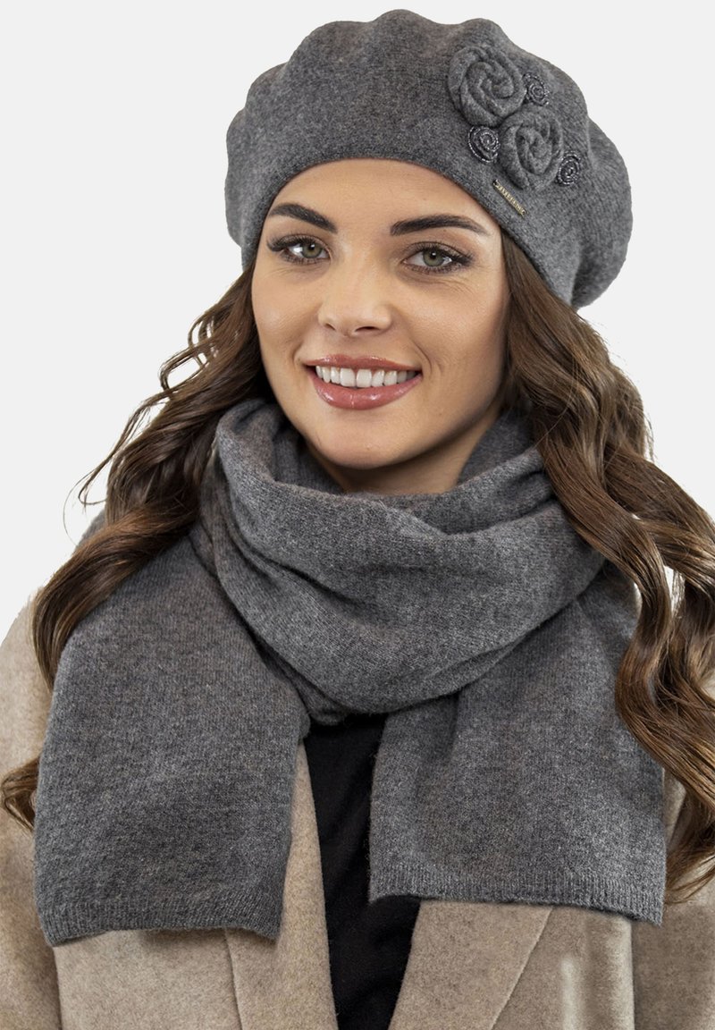 Berretto di lana grigia con accenti floreali e sciarpa grigia, entrambi caratterizzati da una texture morbida. Indossato sopra un cappotto beige, con strati interni neri visibili.