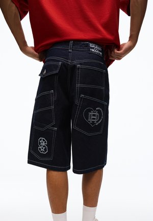 Person trägt schwarze Denim-Shorts mit mehreren großen Taschen und weißen Nähten, kombiniert mit einem roten Hemd und weißen Socken.