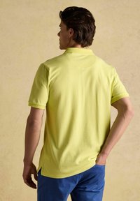 Polo tričko v jasne žltej bavne s krátkymi rukávmi a klasickým golierom. Obsahuje bočné rozparky na leme a hladkú textúru.