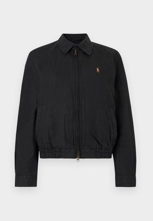 Žena s dlhými tmavými vlasmi, ktorá nosí hnedú čiapku, čiernu mikinu na zips cez svetloružové tričko, biele nohavice a biele tenisky, stojaca s rukami v vreckách.