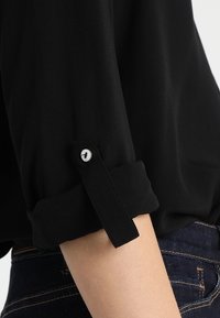 Ærmet af en oprullet sort skjorte, sikret med en knapklap, båret med mørkeblå jeans på en persons arm.