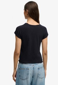 Mujer de cabello largo con una camiseta negra de manga corta y vaqueros de mezclilla azul claro, vista desde atrás sobre un fondo blanco.