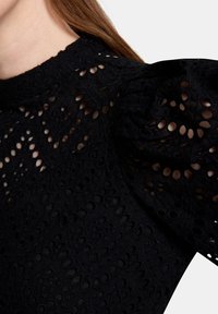 Svart spetsblus med intrikata utskurna mönster, texturerad tyg och hög krage med puffade korta ärmar.