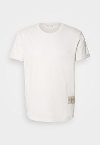 Vit bomullst-shirt med rund halsringning och korta ärmar, med en fyrkantig beige tygetikett med logotyp på vänster sida.