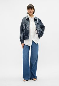 Jūras zila spīdīga ādas jaka ar divām priekšējām kabatām, saskaņota ar baltu pogu krekli un platām gaiši zilām biksēm.