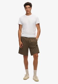 Camiseta blanca de algodón y pantalones cortos de algodón verde oliva. Los pantalones cortos tienen un pequeño logo negro en el dobladillo. El modelo lleva zapatillas beige y calcetines claros.