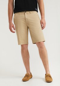 Beige bomullsbyxor med en klassisk passform, med en enkel knappstängning och sidofickor, tillsammans med bruna mockaloafers.