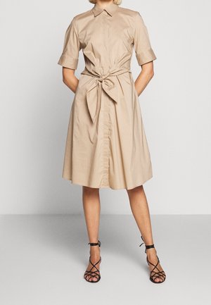 Vrouw in een beige knielang jurkje met korte mouwen en een strik aan de voorkant, gecombineerd met zwarte sandaaltjes met hoge hakken en banden.