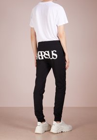 Pantalones de chándal negros con "VERSUS" en grandes letras blancas en la parte posterior, combinados con una camiseta blanca sencilla y zapatillas blancas gruesas.