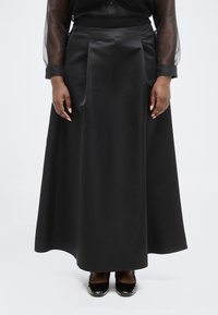 Γυναίκα που φοράει μακριά μαύρη σατέν φούστα, διάφανα μαύρα μανίκια πουκάμισου και μαύρα παπούτσια από δέρμα λουστρίνι, στέκεται μπροστά σε λευκό απλό φόντο.