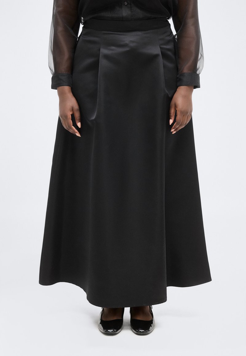 Γυναίκα που φοράει μακριά μαύρη σατέν φούστα, διάφανα μαύρα μανίκια πουκάμισου και μαύρα παπούτσια από δέρμα λουστρίνι, στέκεται μπροστά σε λευκό απλό φόντο.