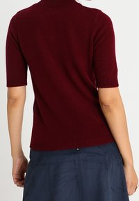 Burgundin värinen lyhythihainen neulepaita, joka on valmistettu pehmeästä neuloksesta ja jossa on muotoiltu siluetti sekä yksityiskohtina ribbaus. Paitaa on yhdistetty tummansiniseen hameeseen.