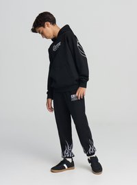 Czarna bluza z kapturem z kieszenią kangurka z przodu i białym nadrukiem na klatce piersiowej, w komplecie z czarnymi dresami ozdobionymi wzorami płomieni na dole nogawek.