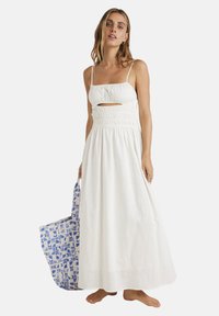 Abito maxi bianco con vita arricciata e dettaglio a taglio. Spalline sottili e tessuto morbido. Una borsa tote blu con motivo viene tenuta in mano.