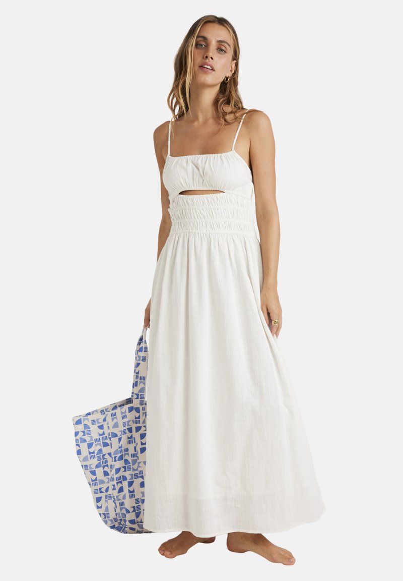Abito maxi bianco con vita arricciata e dettaglio a taglio. Spalline sottili e tessuto morbido. Una borsa tote blu con motivo viene tenuta in mano.