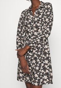 Černé květinové šaty s dlouhými rukávy, zapínáním na knoflíky vpředu a vypasovaným pasem. Mají bílé a oranžové květy na tmavém pozadí.