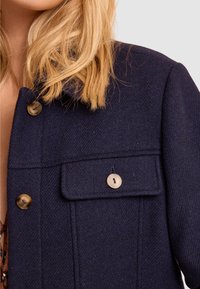 Giacca in misto lana di colore blu navy con design a colletto, bottoni visibili e una tasca sul petto con patta. Tessuto texturizzato con motivo a spina di pesce.