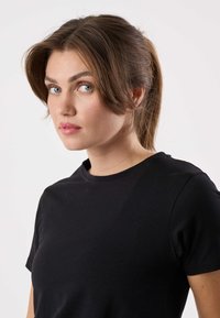 Jonge vrouw met bruin haar in een staart, blauwe ogen en roze lippen, gekleed in een effen zwart t-shirt, kijkt iets zijwaarts tegen een witte achtergrond.