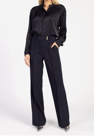 Woman wearing a black satin button-up blouse tucked into high-waisted navy trousers with a matching belt and silver pointed shoes.