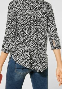 Donna che indossa una blusa a maniche tre quarti con motivo a foglie bianco e nero, pantaloni jeans blu, mentre tiene un piccolo oggetto rosso nella mano sinistra.