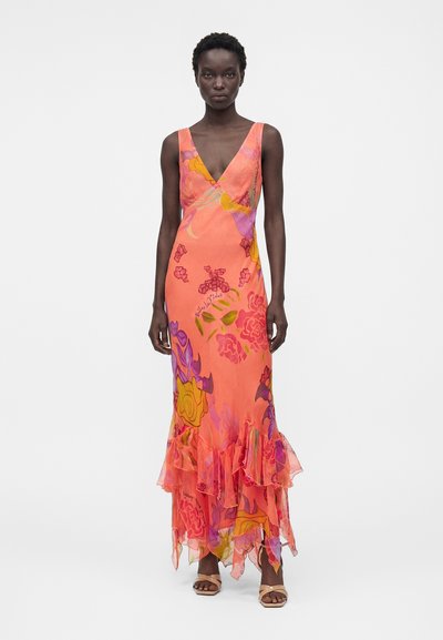 Γυναίκα που φοράει πορτοκαλί μακρύ φόρεμα χωρίς μανίκια, με V-λαιμόκοψη, ροζ, μοβ και κίτρινα λουλουδάτα σχέδια και βολάν στο τελείωμα, σε συνδυασμό με μπεζ σανδάλια.