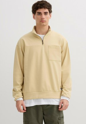 Joven con un pulóver beige de cremallera corta sobre una camisa blanca y pantalones cargo oliva, posando frente a un fondo claro y liso.