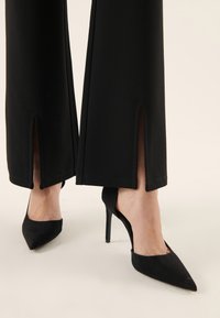 Černé široké kalhoty s postranními rozparky na lemu, v kombinaci s černými vysokými jehlovými botami s špičatou špičkou. Hladká, texturovaná tkanina.