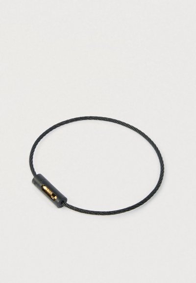 Melnā savērpta tērauda kabelis cilpa ar mazu melnu un zeltītu kombināciju atslēgu uz gaiša fona.