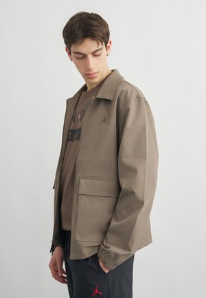 Jaunas vyras žvelgia žemyn, vilkėdamas rudą striukę, derančią rudą marškinius ir juodas kelnes su raudonu logotipu, stovėdamas ant vienspalvio šviesaus fono.