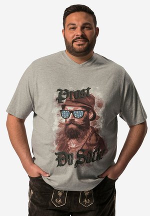 Uomo con capelli corti e scuri e barba indossa una maglietta grigio chiaro con grafica di uomo barbuto con occhiali da sole e pantaloni scuri con ricamo, mani in tasca.